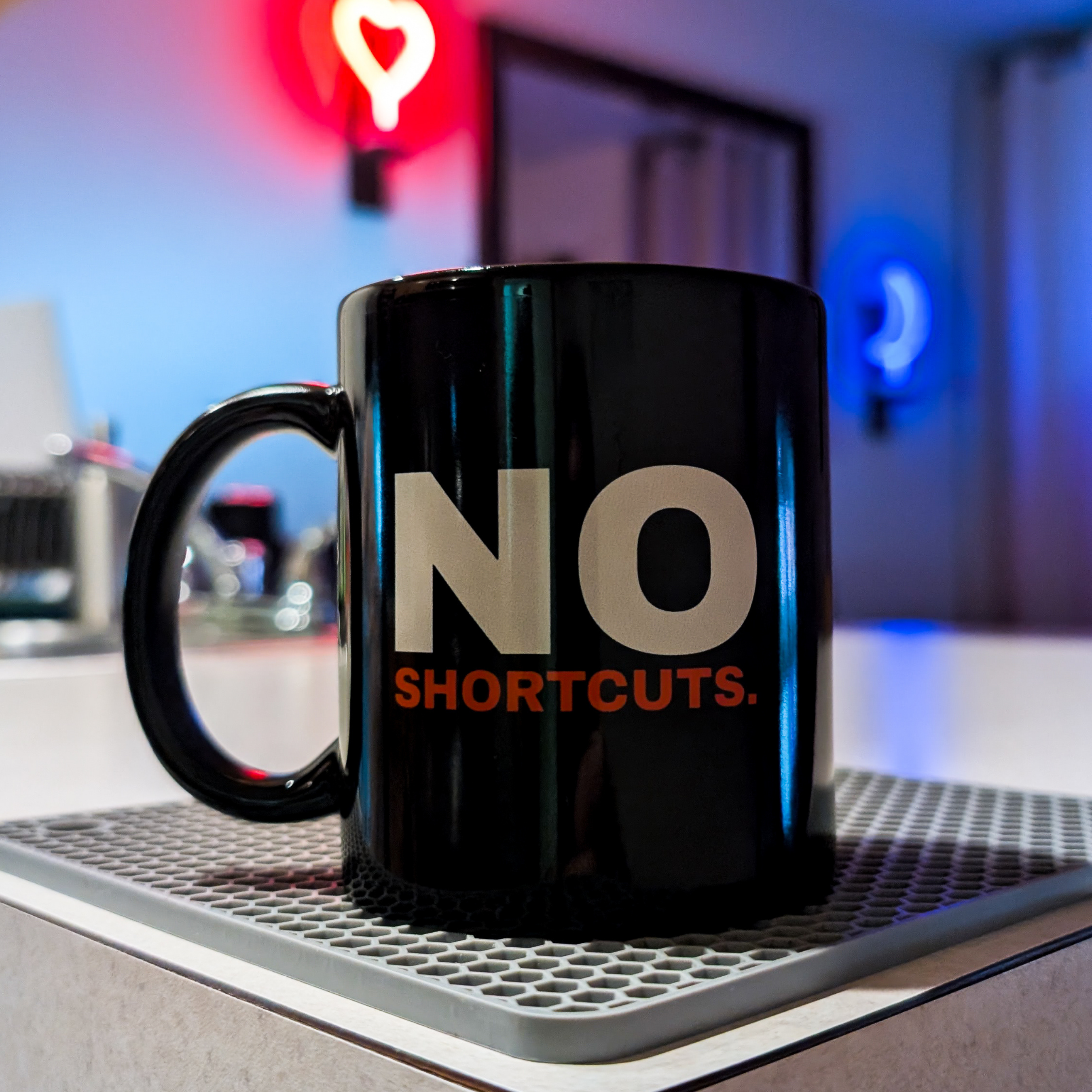 Black mug with 'NO SHORTCUTS.' text on a metallic surface with a blurred background