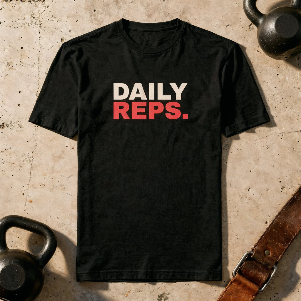 Black t-shirt with 'DAILY REPS.' text on a textured surface with kettlebells and a belt.